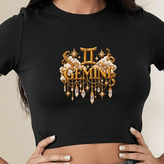 A woman wearing a black short-sleeve crop top with a gold and white gemstone-themed graphic that reads 'II GEMINI' on the chest, paired with blue jeans in a bright indoor room.